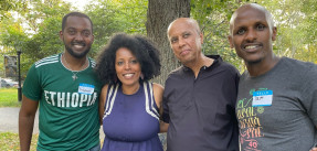 Four smiling black people, three men and one woman, with arms linked around one another