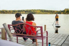 A couple sitting on a river deck bench taking a picture of their son on a paddle surfboard