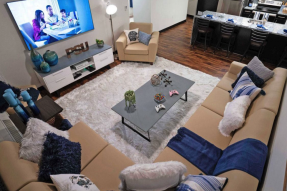 A Staged dorm living room with couches, TV, coffee table, white rug, and kitchen in the background