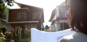 A woman reviewing house blueprints looks to her left from the right of frame while standing in front of new homes.