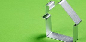 A picture of a silver cookie cutter in the shape of a house on a bright green background.