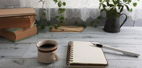 A picture of an open journal on a light-grey desk with a pen resting on it, next to a filled coffee mug, books, and a plant.