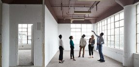 A group of people touring an office building point to a ceiling with exposed pipes.
