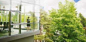 A woman stands in a commercial building with walls of glass windows looking out to trees neighboring the building.