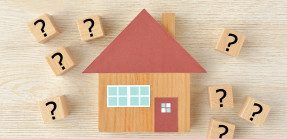 A picture of a 2D wooden house miniature with question marks painted on small wood squares surrounding it.