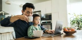Father multitasking at computer with child in lap.