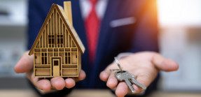 A picture of a man in a suit holding a house miniature in one hand and a set of keys in the other