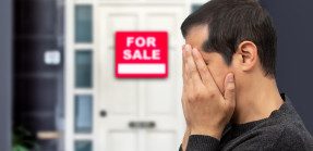 Man holding head in hands in front of for sale sign on house