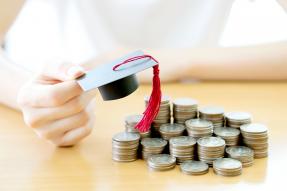 Person holding a tiny mortarboard over stacks of coins