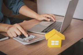 Person typing on laptop and calculator next to a yellow model scale home