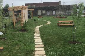 A park with a walking path, picnic tables, recently planted trees, and trellises.