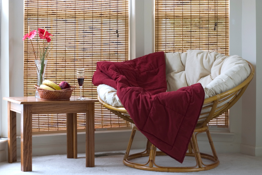 papasan chair near coffee table, which has wine, flowers and a bowl of fruit on it. Windows are covered in bamboo shades.