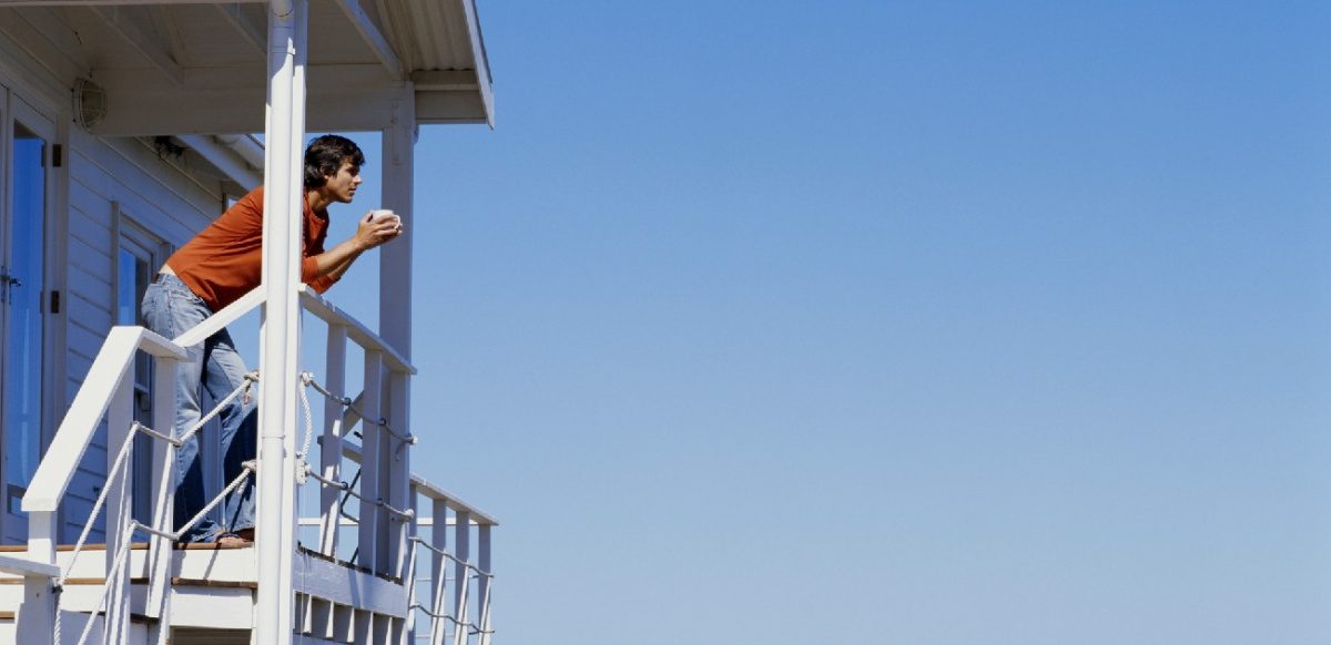 A person stands on the balcony of their vacation home with a cup of coffee, looking out across the view.