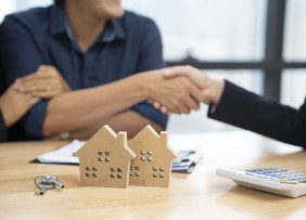 Couple shaking hands with business professional over a desk with a contract, a key, small wooden houses, and a calculator