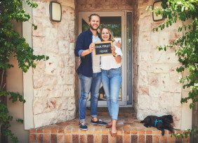 Young couple standing in front of their new home holding a chalkboard with "first home" written on it
