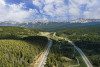 Aerial view of highway through trees with mountains, clouds, and blue sky